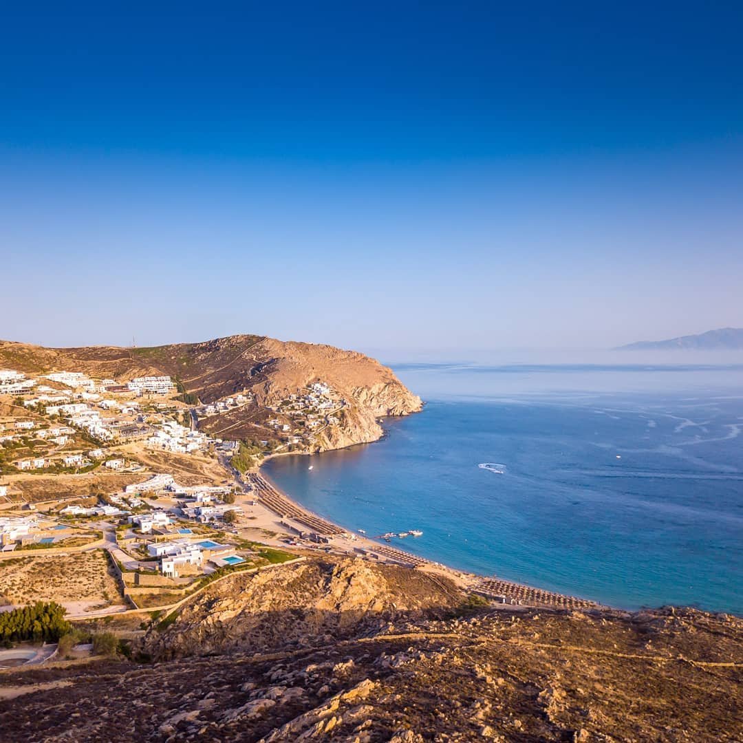 Pristine Mykonos beach on the south coast, featuring calm, shallow waters and scenic cliffs in the background.