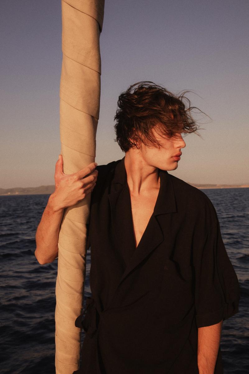 Man Standing on the Bow of a Sailing Yacht – "A stylish man standing at the bow of a yacht, looking out at the endless sea, with the sun setting behind him.